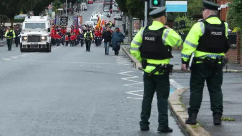 Pacemaker Police in the forground with their back to the camera. In the background a parade of people in red uniform led by a police land rover