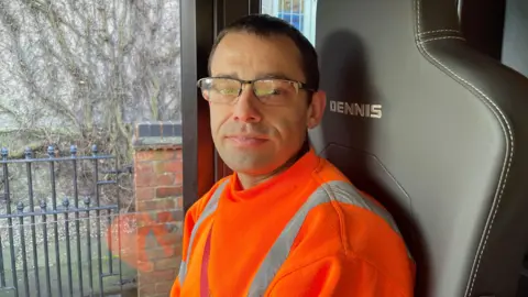 A man in the cab of a bin lorry. He is wearing glasses and has short hair. We can see a seat branded "Dennis" behind him. He is smiling and wearing a orange hi-vis top.