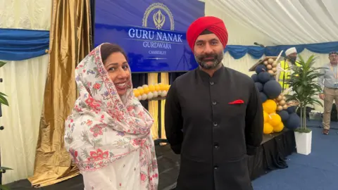 BBC/Julia Moore Woman with a flowery headscarf and cream dress, standing alongside a man in a black suit with red hankerchief and red turban.