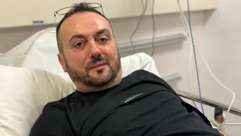 Zoran Gucev is lying on a hospital bed. He has dark hair and beard and has glasses balanced on his forehead. He is wearing a dark top. 