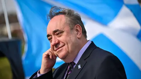 Alex Salmond wears a dark suit with a lilac shirt and a patterned black and purple tie. He stands in front of a saltire.