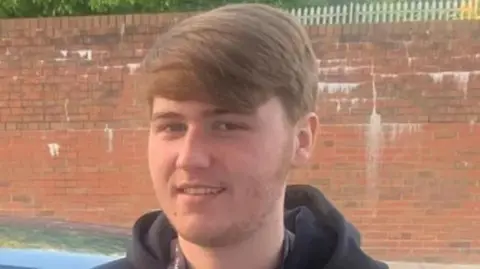 Ellis Cox in front of brick wall. He is smiling and wearing a black hooded jumper. He has light brown hair and a small amount of facial hair.