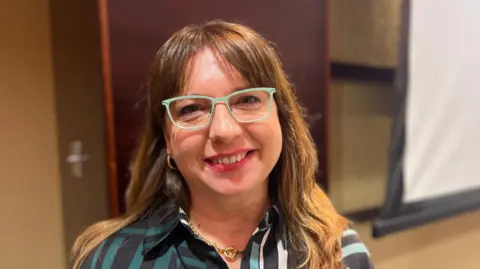 Ana is a woman with brown hair. She is wearing green glasses and a green and black dress with a gold necklace. She is stood in a conference room and is smiling at the camera.