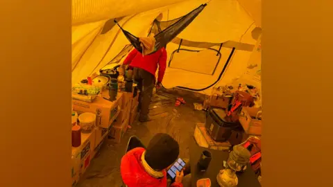 BAS Two people are in a tent. They are wearing jackets and woolly hats and are surrounded by boxes of equipment and provisions including cooking equipment