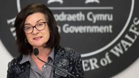 Getty Images A woman with dark hair, wearing dark glasses, a grey shirt and a black and grey blazer. She is speaking into a microphone in front of a Welsh government sign