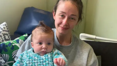 Laura Derrick holding her young baby indoors, seated on a sofa with cushions visible in the background.