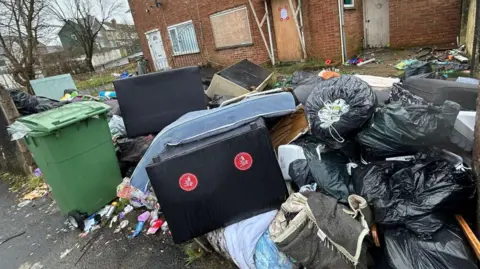 A bed frame and mattress, a sofa and multiple bin bags are piled up in the garden of a house. A green wheelie bin is also placed on the pavement outside the house. The window and front door of the property are boarded up.