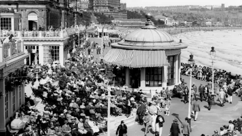North Yorkshire Archives Scarborough Spa pictured in the early–mid 20th century, captured in black and white, with large crowds. Mid-picture is a round pavilion with a domed roof and striped awning. In the background on a hillside there is a series of large buildings and hotels overlooking the bay.