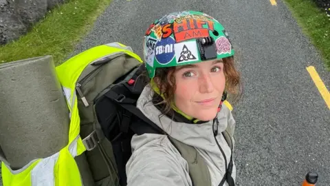 Becky Gilmour Una mujer con cabello largo y castaño y ojos marrones mira a la cámara para tomarse una selfie. Lleva puesto un casco de ciclista verde, decorado con pegatinas de colores brillantes. Lleva una mochila en la espalda, parcialmente cubierta por una chaqueta de alta visibilidad.