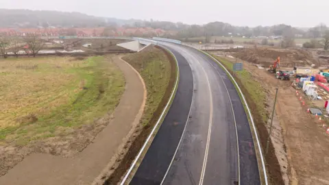 Chester Bridge in Ash Road. The aerial photo of the bridge shows a road leading to the bridge with a construction site on the right hand site. The construction site has tractors and diggers in it.