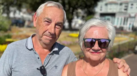 Stuart Woodward/BBC John and Sue Wackett standing by the seafront in Clacton. They both have grey hair. Sue is wearing a brown vest top and sunglasses. John is wearing a grey polo top and squinting due to the sun.