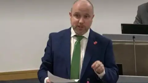 Senedd Cymru Mabon is standing in the chamber holding a paper in one hand and gesticulating with the other. He is wearing a blue suit and green tie. He is bald on top and has a cropped beard. 