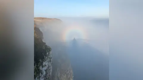 Chris Atkinson An image of a person's shadow, encased in a rainbow, off Bempton Cliffs in East Yorkshire. It is a sunny day with a fret coming from the sea