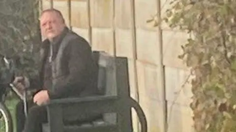 A grainy picture of a man dressed in black trousers, a black jumper and a puffer jacket sitting on a park bench with a bicycle in the background.