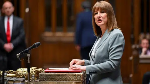 Reuters Chancellor Rachel Reeves standing up in parliament. She has mid length brown hair and a fringe and is waring a grey suit jacket and white t-shirt underneath.