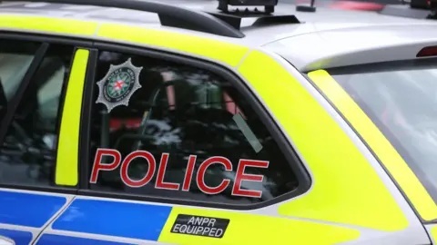 A yellow and blue police car with 'police' written on the back black tinted windows. A police badge is also on the window. The roof is grey with black roof racks.