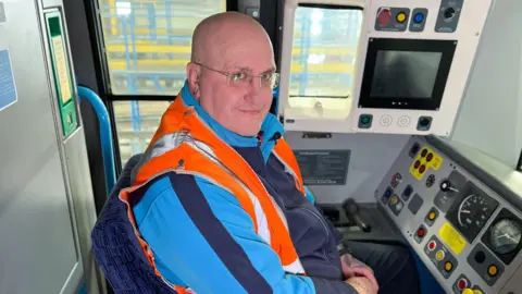BBC Train driver David Boffin sitting in a train cab wearing a hi vis jacket and looking to camera.