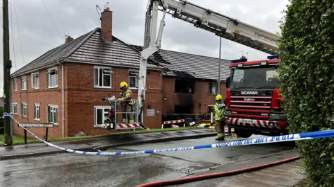 BBC Police tape is cordoning off a road. There is a fire engine with an aerial platform parked next to a block of two-storey flats. A section of the flats is black with smoke damage and there are roof tiles missing. There are two fire officers, one in the aerial lift, and one standing by the fire engine