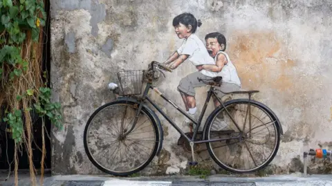 Ernest Zacharevic A photograph of a wall mural, depicting two children in white tops. The images in painted to show the children seated on a real-life bicycle.