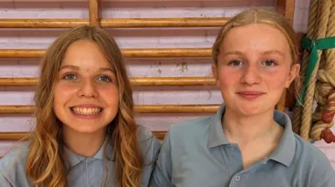 Myfi and Elin sit next to each other smiling at camera. Myfi, on the left, has curly blonde hair. Elin has blonde hair in a low ponytail. Both wears light blue uniform polo tops.