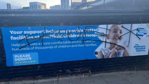 A billboard for the Sheffield Children's Hospital Charity which reads: "Yours support helps us provide better facilities at the hospital." However, a graffiti tag has been sprayed across the entire sign, including a photo of a little girl using a breathing tube.