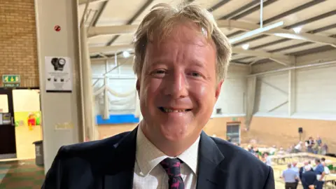 Ben Schofield/BBC Paul Bristow has sandy coloured short hair. He is wearing a dark jacket with white checked shirt, and a purple and black tie. Behind him is a sports hall with an election count taking place.