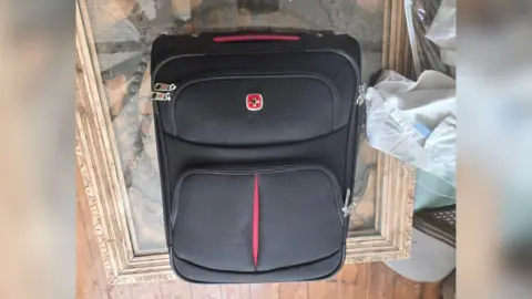 Laura Bayliss Picture of a black suitcase with a red handled and a red logo with a silver cross.