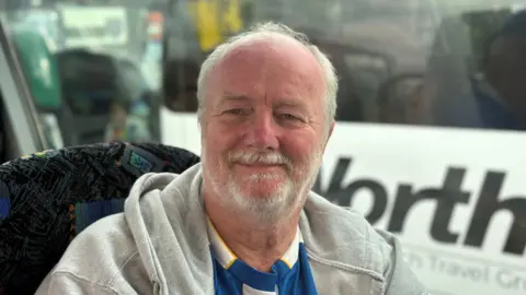 A man with short white hair and facial hair wearing a blue and white Albion shirt, with a grey hoodie on top. He is sat in a coach seat next to the window while smiling at the camera