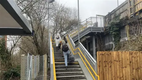 Adrian Harms / BBC Two people walking over up stairs towards a railway station platform.