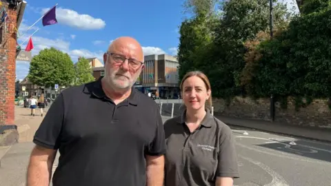 Holly Nichols/BBC Robert and Kim Greenaway, standing outside, both wearing black T-shirts, looking at the camera. There are a number of bollards behind them, shops and people in the distance. There are blue skies on a sunny day.