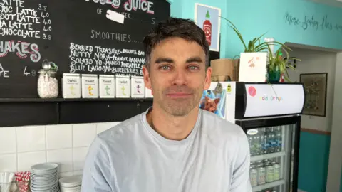 Steve Piper stands behind the counter of his cafe in Shepton Mallet He has short, dark hair and is wearing a light grey T-shirt. He has blue eyes and stubble and he is just managing a small smile.