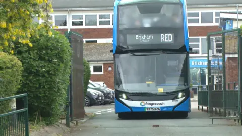 A blue double-decker bus with the Stagecoach logo on the front passes through the gates of a college car park.