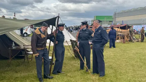 Three men and a woman, wearing air cadet uniforms, with one also wearing a brown bomber jacket, stand talking outside an old-fashioned green tent in the middle of an airfield. More tents and people can be seen behind them.