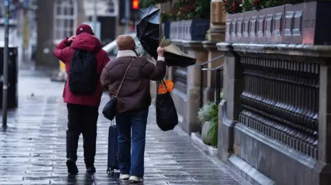 PA Media A man and woman pictured from behind walking down a damp street. The woman is wearing a brown coat and hat, while clutching onto an umbrella which has been blown inside out.