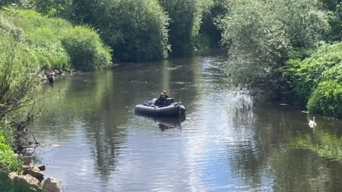 BBC/Phil Bodmer Two people in a rigid inflatable boat in the middle of a river flanked by green foliage.