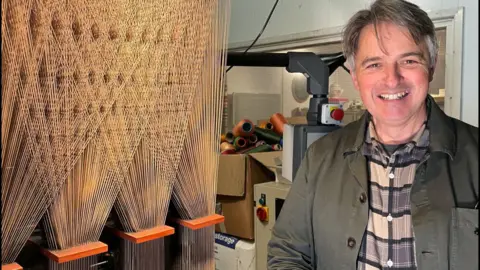 Robert smiles next to the loom which is threaded with silk.