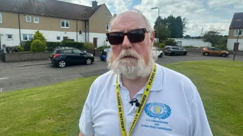 Bill Mackie, 71, with short white hair and a white bears, wears dark glasses for his eye condition and a military polo shirt. He wears a lanyard saying "please be patient, visually impaired". The houses of the estate lie behind him on two sides.