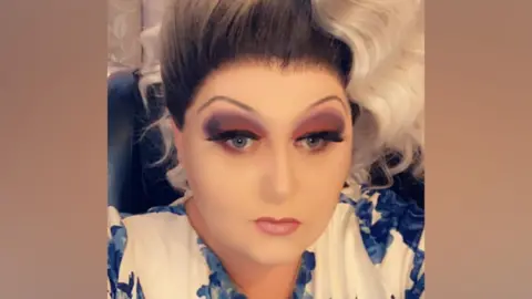 Facebook Scott Hallas aka Dragalicious looks at the camera. He is wearing a blue and white dress and has a heavily made-up face. He is wearing a curly black and white wig.