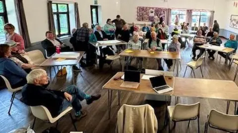 LDRS A room full of desks and chairs. There are people sat down and some stood. Some have laptops and notepads. The room has windows and white walls, it is sunny outside.