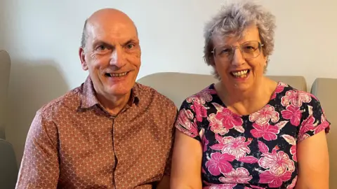 An elderly man with a brown shirt smiles as he sits on a sofa next to an elderly woman wearing a black and pink floral top, who is also smiling.