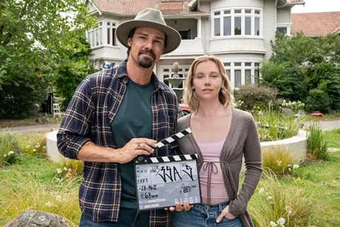 BBC/The Ridge Production Ltd A behind-the-scenes moment from The Ridge shows Lauren Lyle and Jay Ryan standing in front of a large white house with a red-tiled roof and multiple windows. Ryan wears a hat, a checked shirt over a green t-shirt, and holding a clapperboard used in film production. Lauren Lyle is dressed in a light purple top, an open cardigan, and jeans.