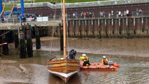 A historic wooden fishing boat is stuck on a sandbank, an orange lifeboat with two crew helps people off the boat