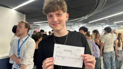 Emy Moore/BBC Adam Thomas Clark smiling into the camera while holding up a folded A4 sheet of paper with his results on which cannot be made out. He has brown, curly hair which is shaved on the sides. He is wearing a black t-shirt and a long necklace and rings on both hands. There is a group of people standing behind him talking in smaller groups. They are in a modern looking room with white walls.