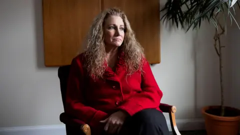Marlean Ames sits in a chair in her lawyer's office. She is gazes off screen and is wearing a red jacket and black pants. Her hands are folded in her lap.