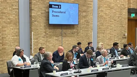 Amy Holmes/BBC A group of officials sit at desks in a council-style meeting room, with papers, microphones, and a screen displaying “Item 3: Procedural Items” on a brick wall behind.