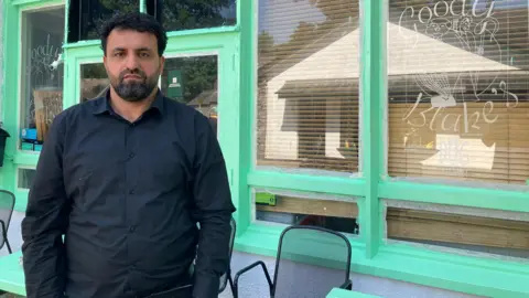 Shwan Khder is wearing a black shirt. He has a beard and looks unhappy. Behind him there is a building with large windows and light green window frames.