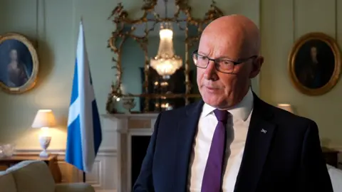 John Swinney wears glasses, a dark suit, white shirt and purple tie