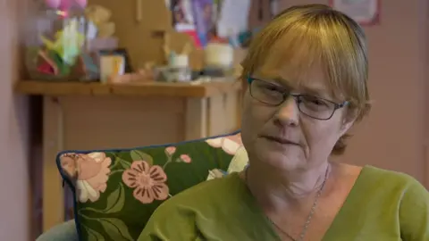 Katherine Elliot who is wearing blue glasses, she has short ginger hair, with a green shirt, sitting behind a green and pink floral cushion.