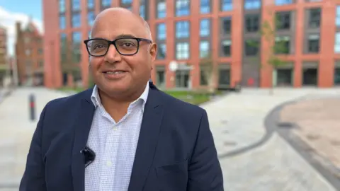 Man stands infront of block of apartments wearing glasses, blue blazer blue shirt