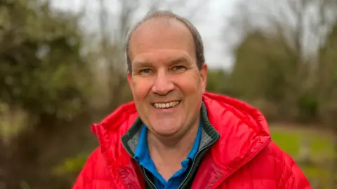 MARTIN GILES/BBC A man smiling at the camera wearing a red puffy jacket in a park, which is out of focus in the background.